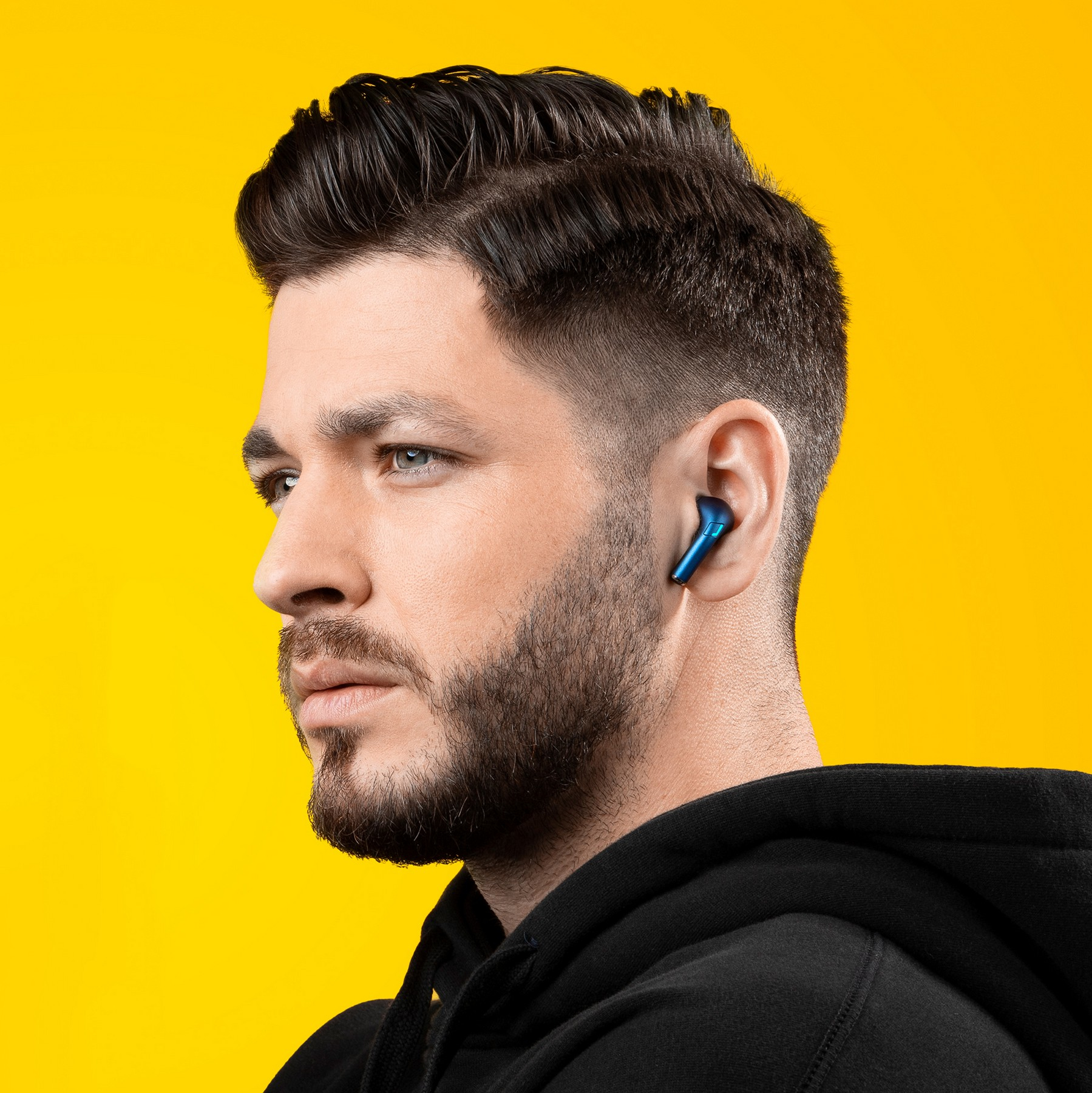 Ein Mann mit dunklen Haaren, Bart und schwarzem Hoodie trägt einen blauen Ohrhörer vor gelbem Hintergrund.
