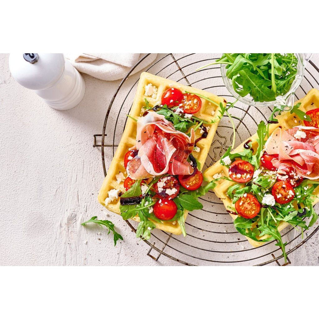 Waffle salati con prosciutto, pomodorini, rucola, formaggio e glassa balsamica su una griglia.