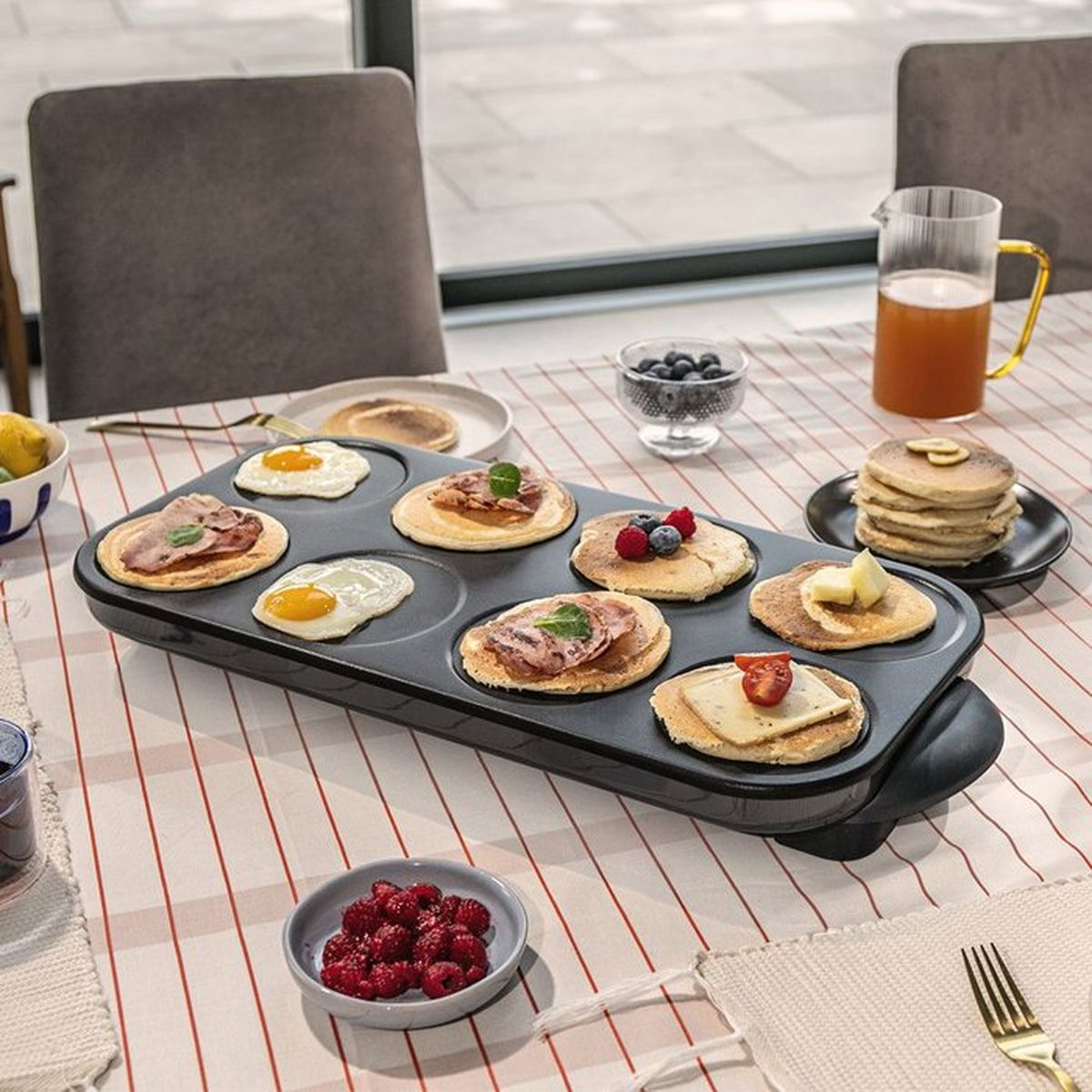 Una piastra elettrica su un tavolo con pancake, uova fritte e bacche.