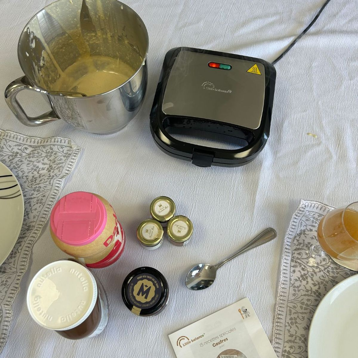 Piastra per waffle, ciotola con impasto, barattoli di crema e cucchiaio su un tavolo bianco.