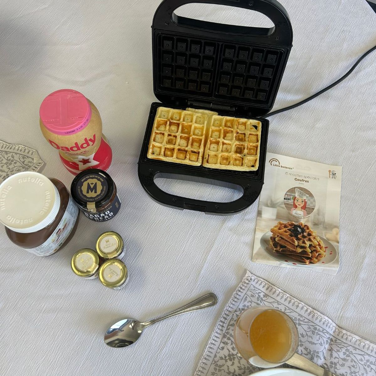 Una piastra per waffle con waffle freschi, creme, ricette e un bicchiere di succo su un tavolo.