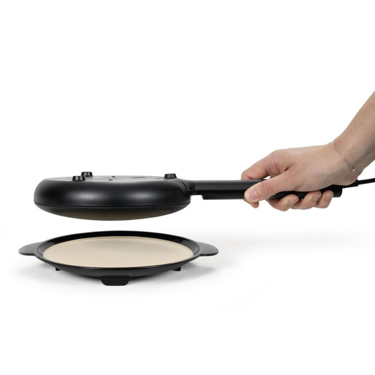 Una mano tiene una piastra elettrica nera sopra la sua base riscaldante beige e rotonda su sfondo bianco.