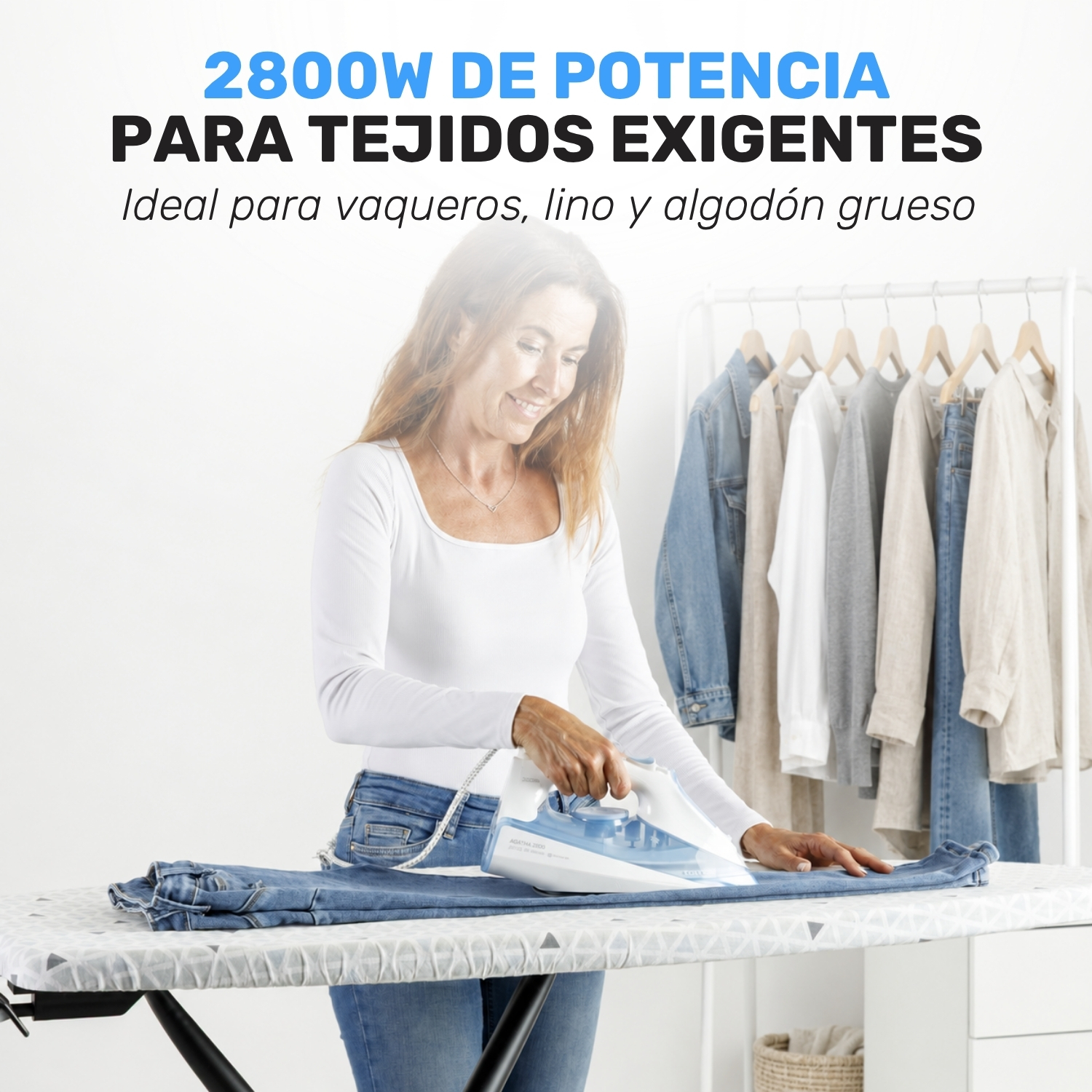 Una mujer plancha unos vaqueros en una tabla de planchar con ropa colgada de fondo.
