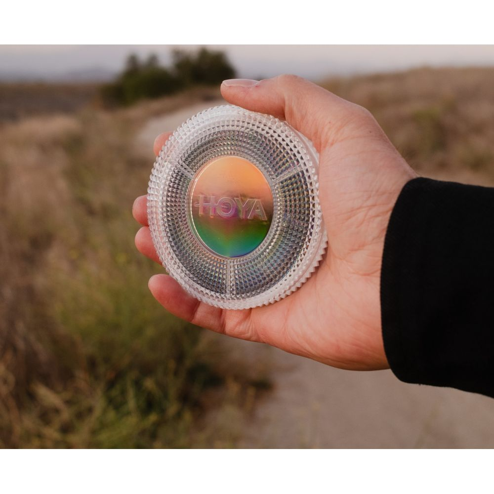Eine Hand hält einen runden, strukturierten Glasfilter mit dem Hoya-Logo vor verschwommenem Hintergrund.