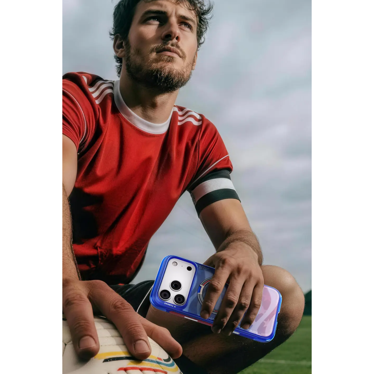 Kırmızı formalı bir futbolcu çimlerde diz çökmüş, elinde akıllı telefon ve top tutuyor.