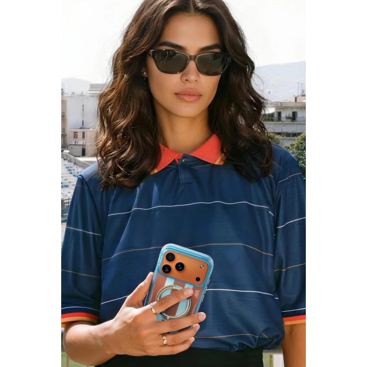 Güneş gözlüklü ve mavi polo yaka tişörtlü esmer bir kadın, arka planda şehirle birlikte telefon tutuyor.