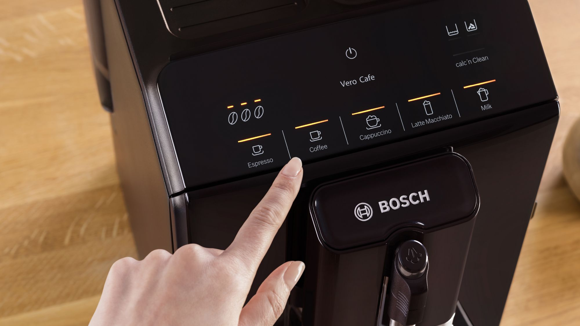 Una mano pulsa la pantalla táctil iluminada de una cafetera Bosch Vero Cafe negra sobre una mesa de madera.