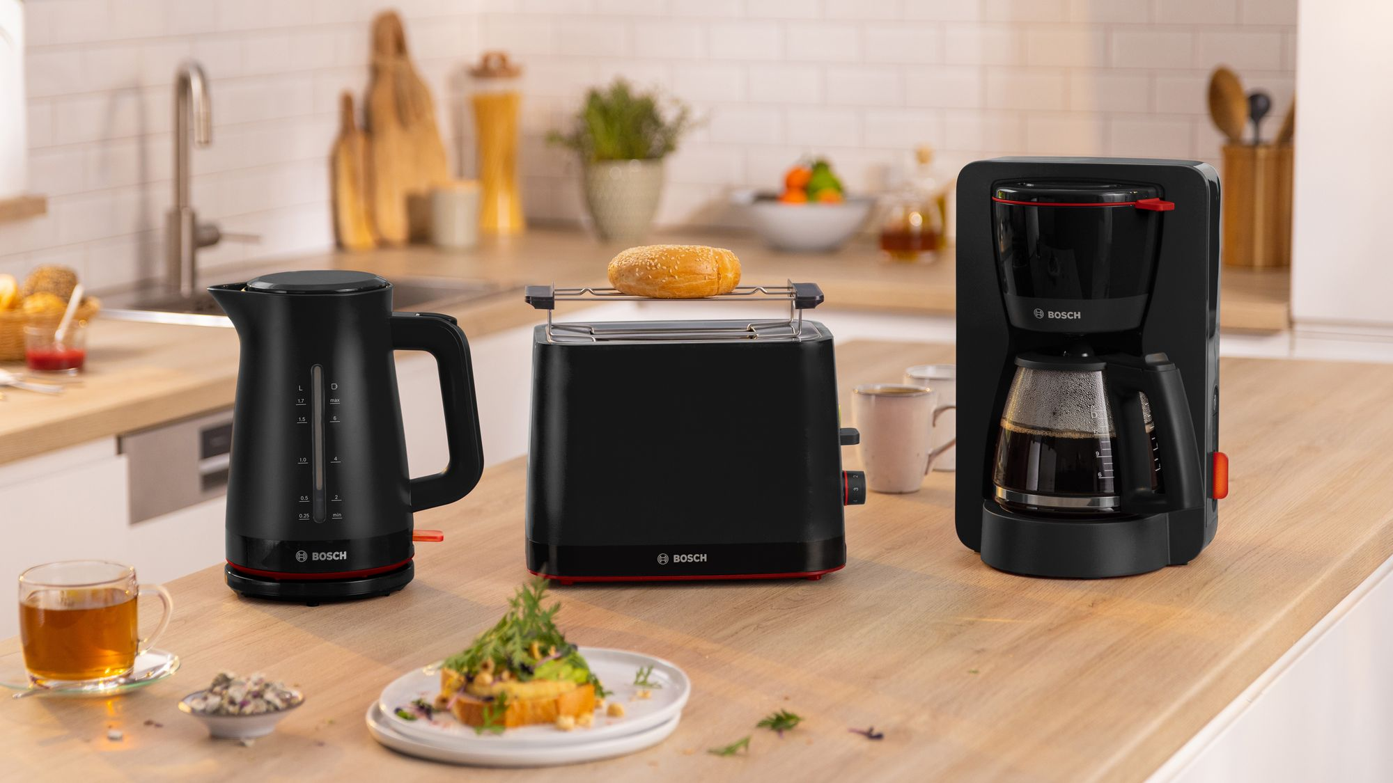 Electrodomésticos Bosch negros en encimera de madera, incluyendo hervidor, tostadora con pan y cafetera.