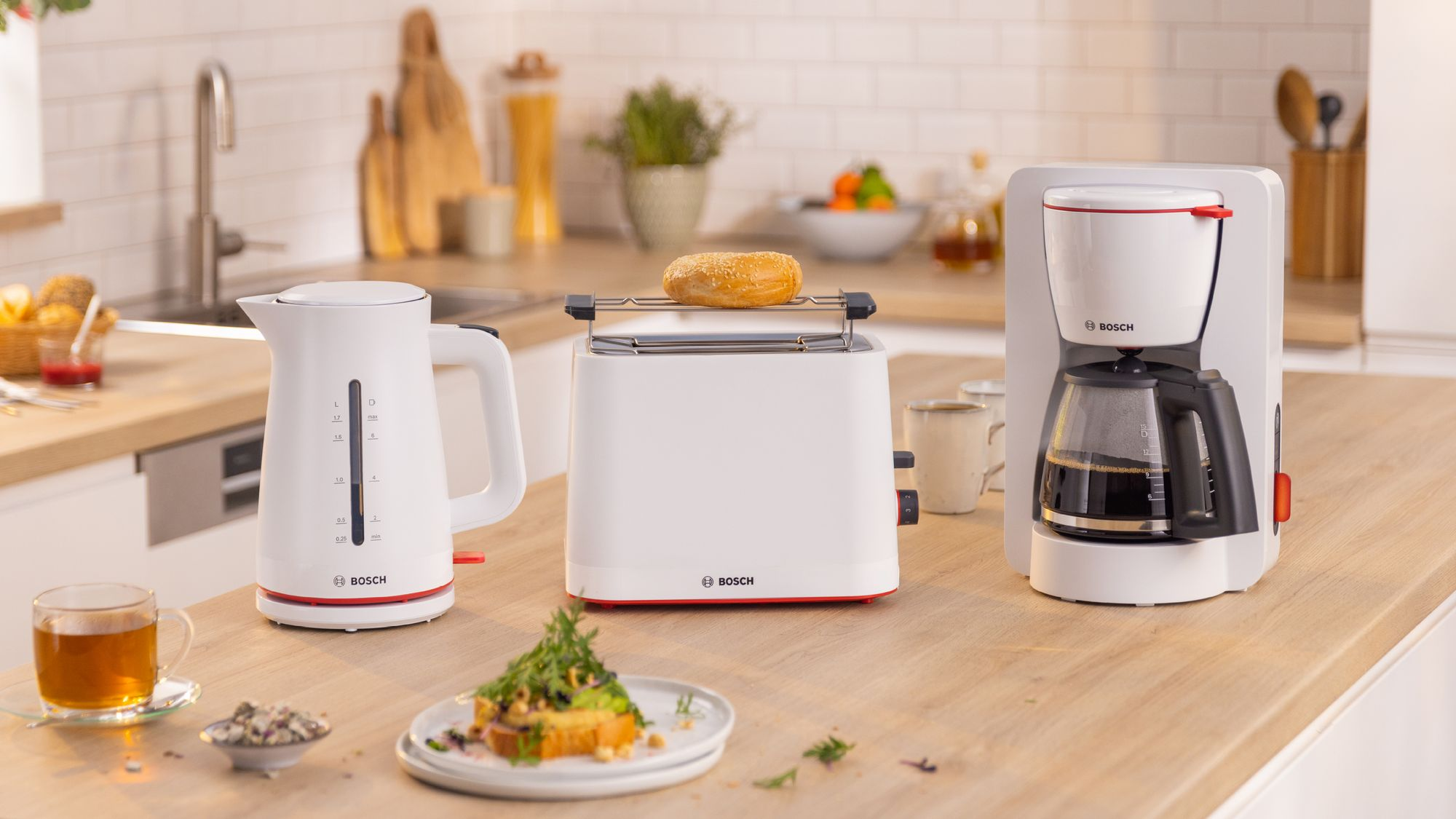 Electrodomésticos blancos Bosch como hervidor, tostadora con panecillo y cafetera sobre una encimera de madera.
