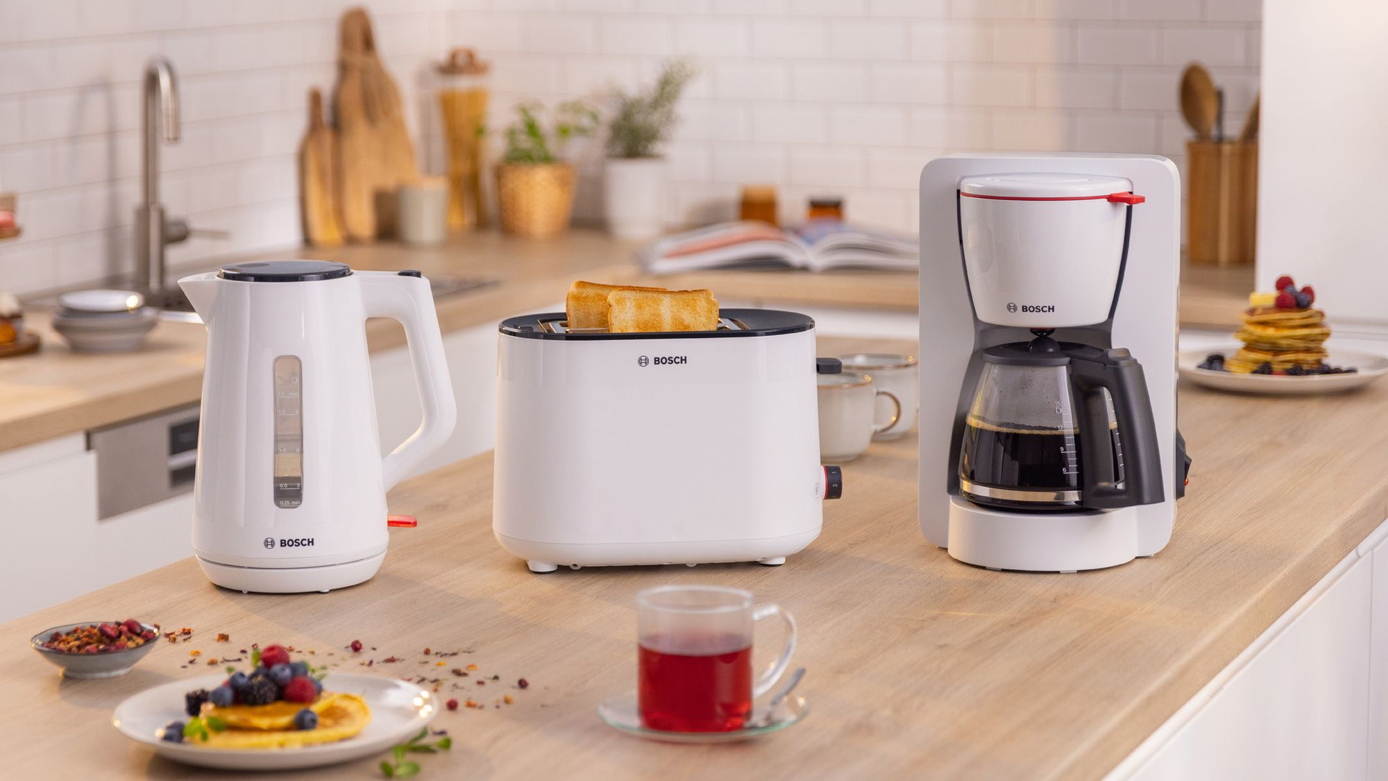 Electrodomésticos blancos Bosch como hervidor, tostadora con tostada y cafetera en una encimera de madera.