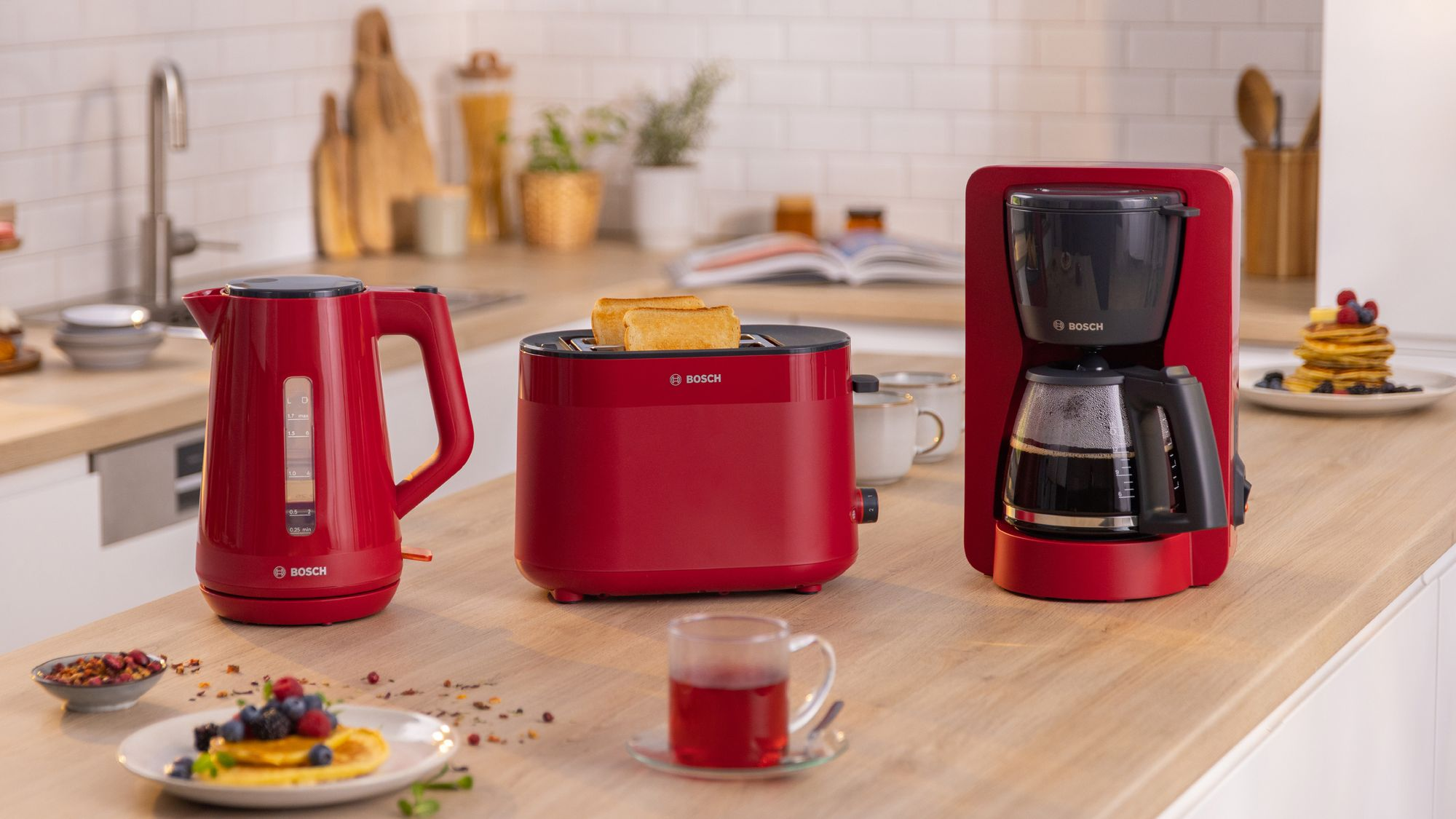 Un juego de electrodomésticos de cocina Bosch rojos con hervidor, tostadora con tostadas y cafetera sobre encimera.
