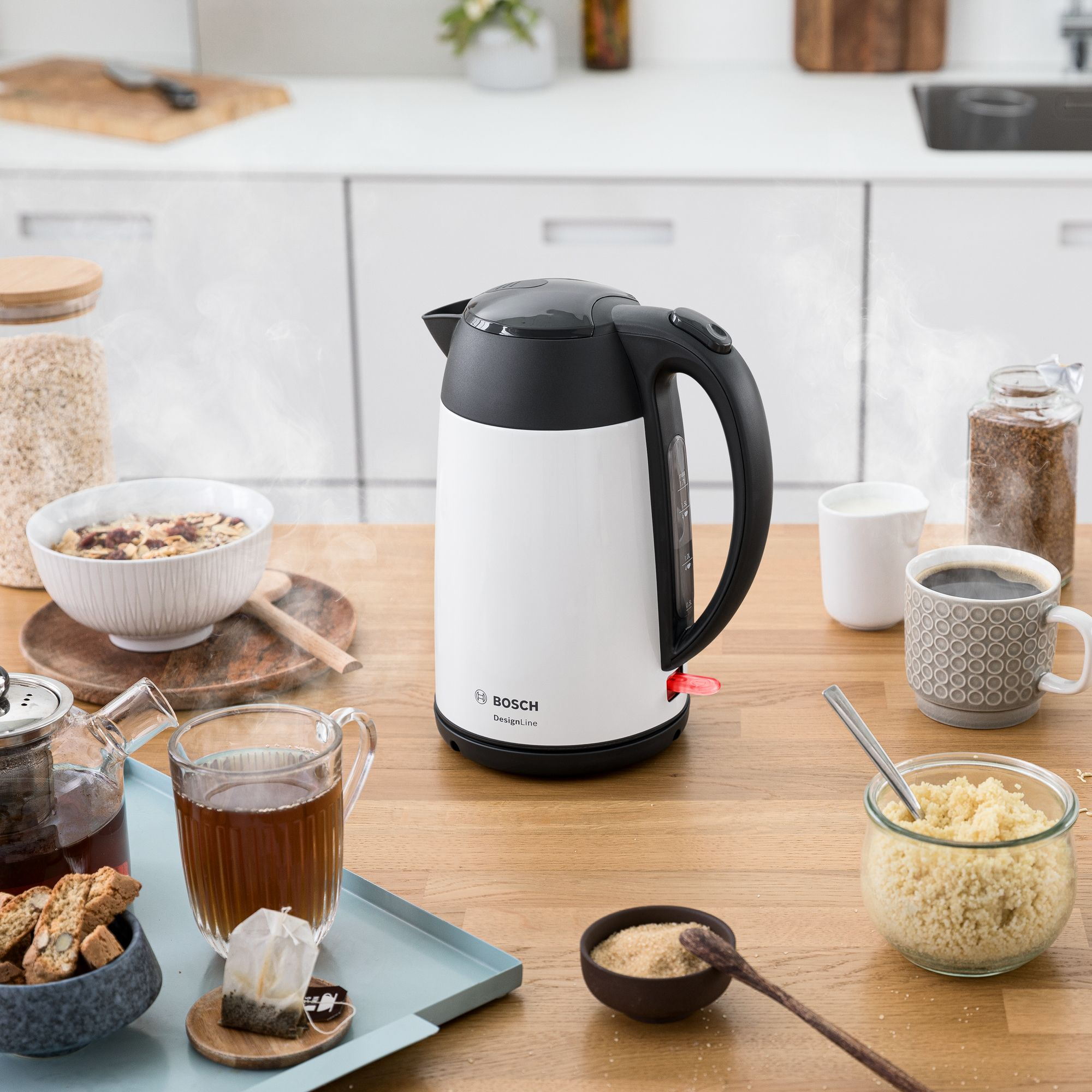 Un hervidor eléctrico Bosch blanco y negro en una mesa de madera con desayuno, vapor y cocina difuminada.