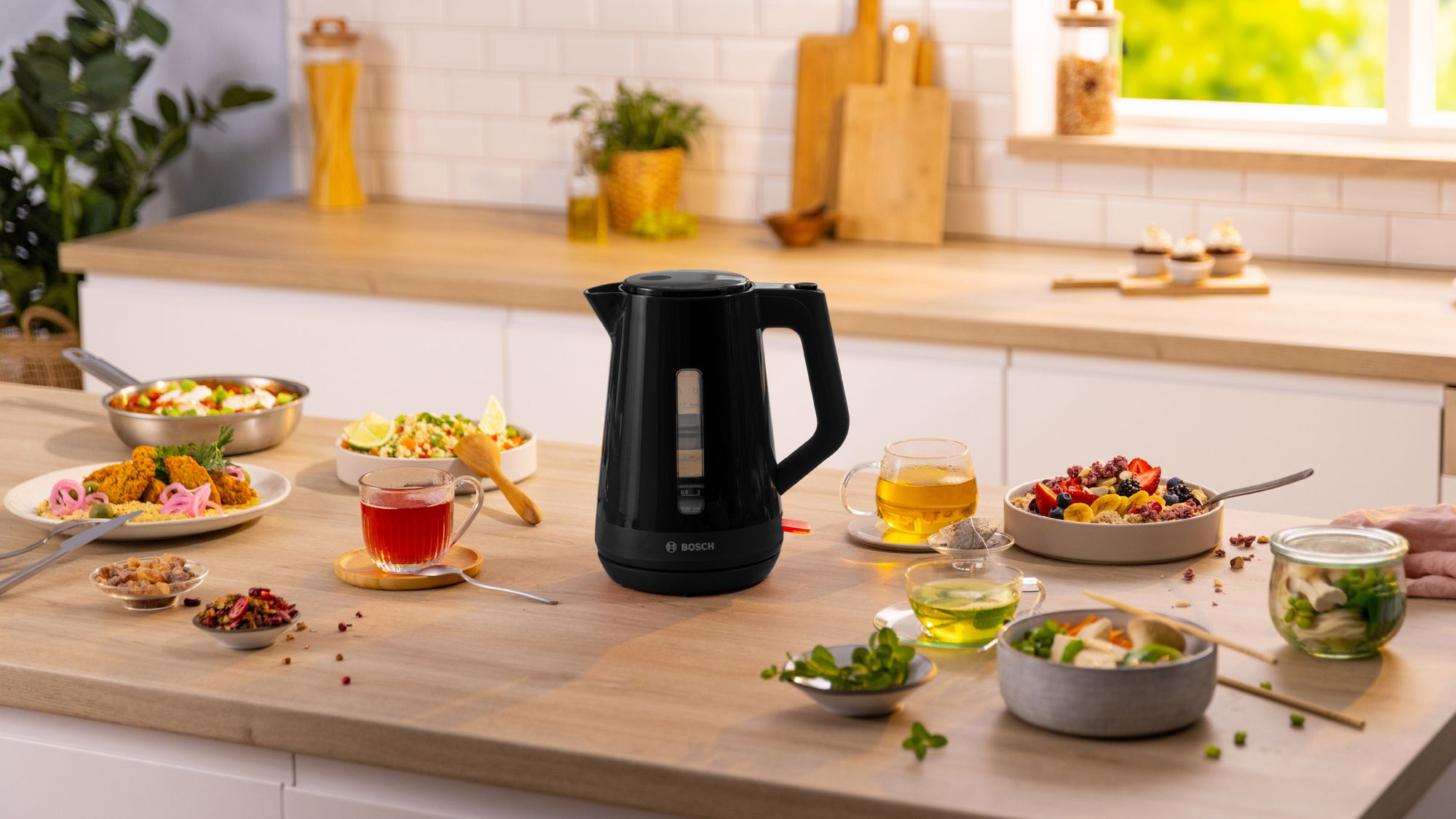 Un hervidor eléctrico negro Bosch descansa sobre la encimera de una cocina con varios platos y bebidas.