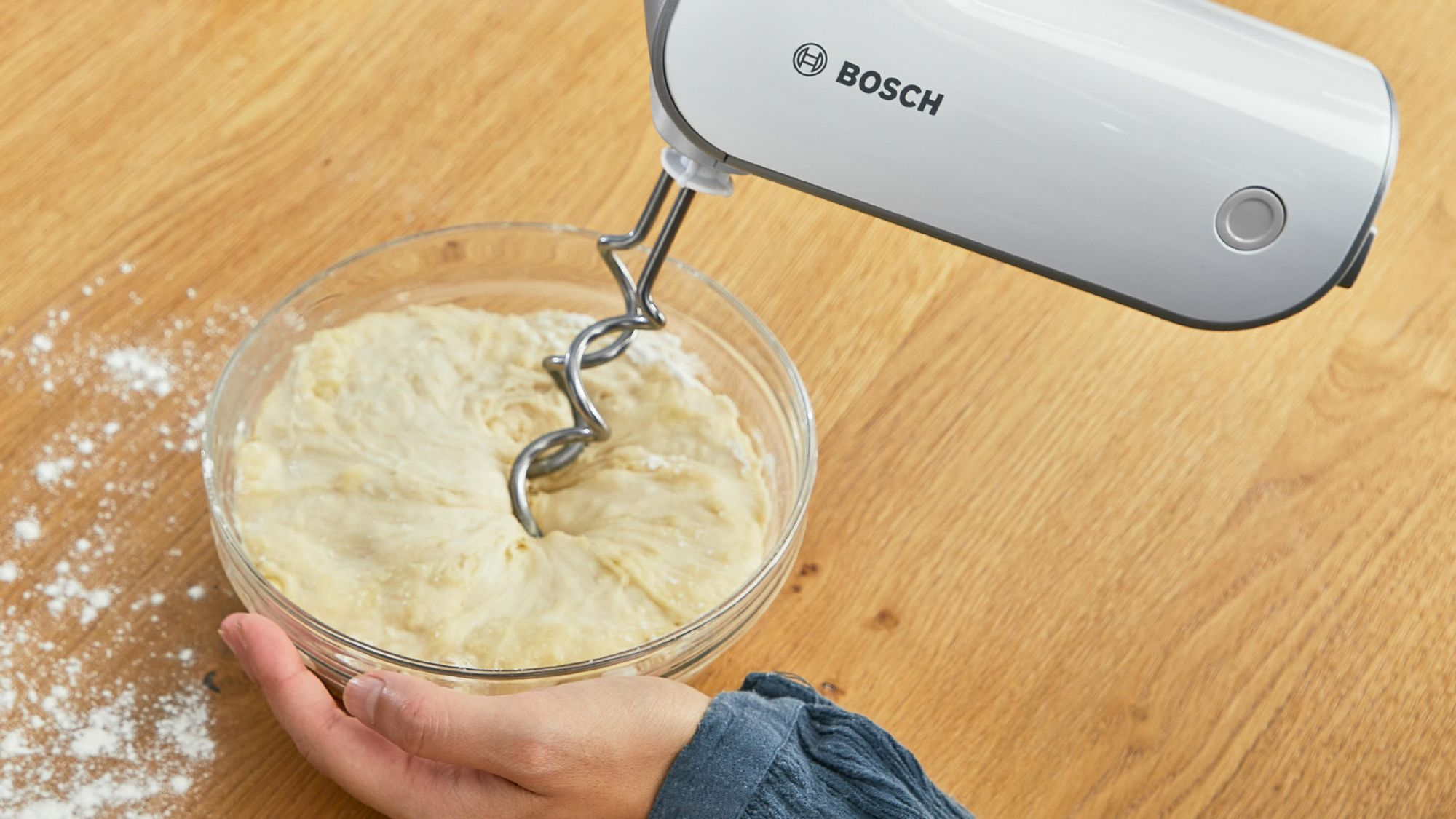 Una batidora de mano Bosch blanca amasa masa en un bol de cristal sobre una mesa de madera.