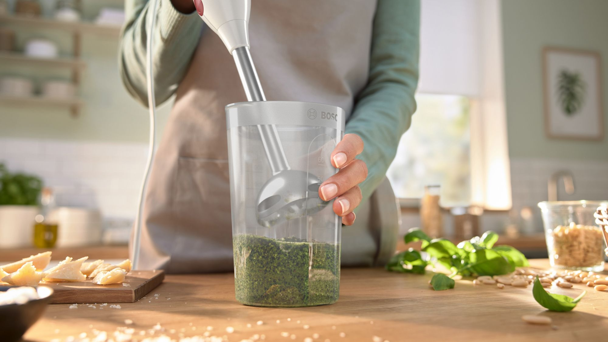 Una persona usa una batidora de mano en un vaso de plástico con mezcla de hierbas verdes en una cocina.