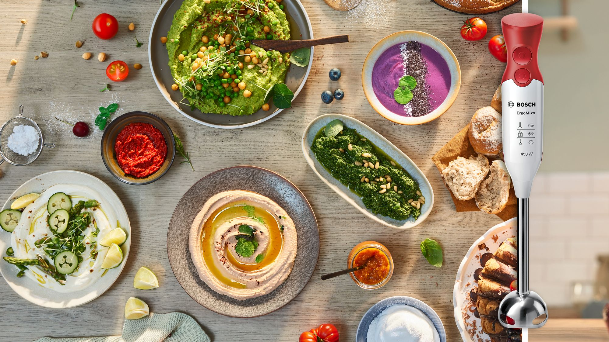 Una batidora de mano roja y blanca se encuentra entre tazones de hummus, salsas verdes y sopa morada en una mesa.