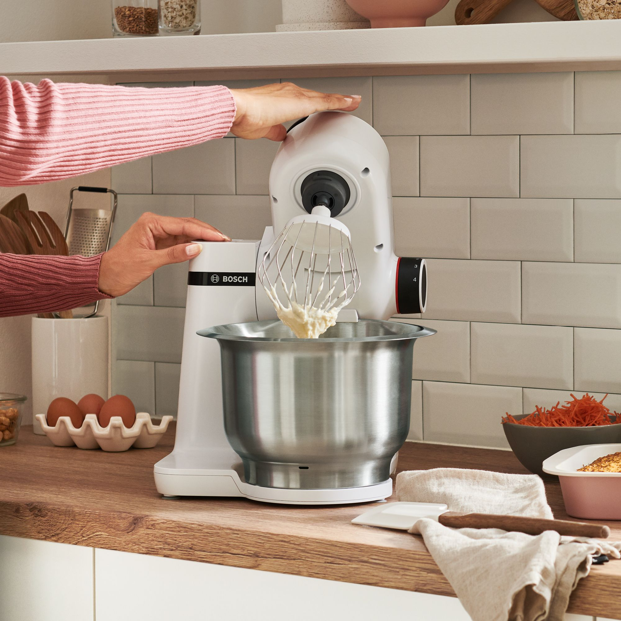 Persona operando un robot de cocina Bosch blanco con bol de acero inoxidable en una encimera de madera.