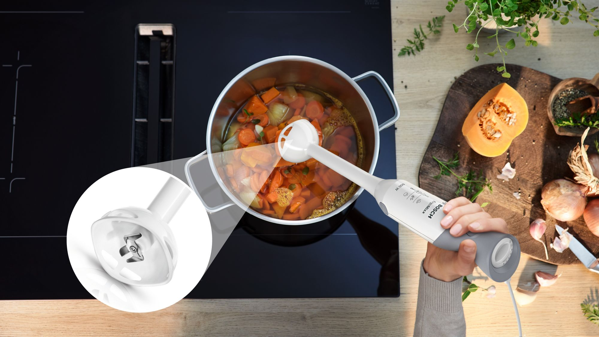 Ein weißer Stabmixer püriert Suppe in einem Topf auf einem Kochfeld, mit vergrößertem Detail der Klinge.