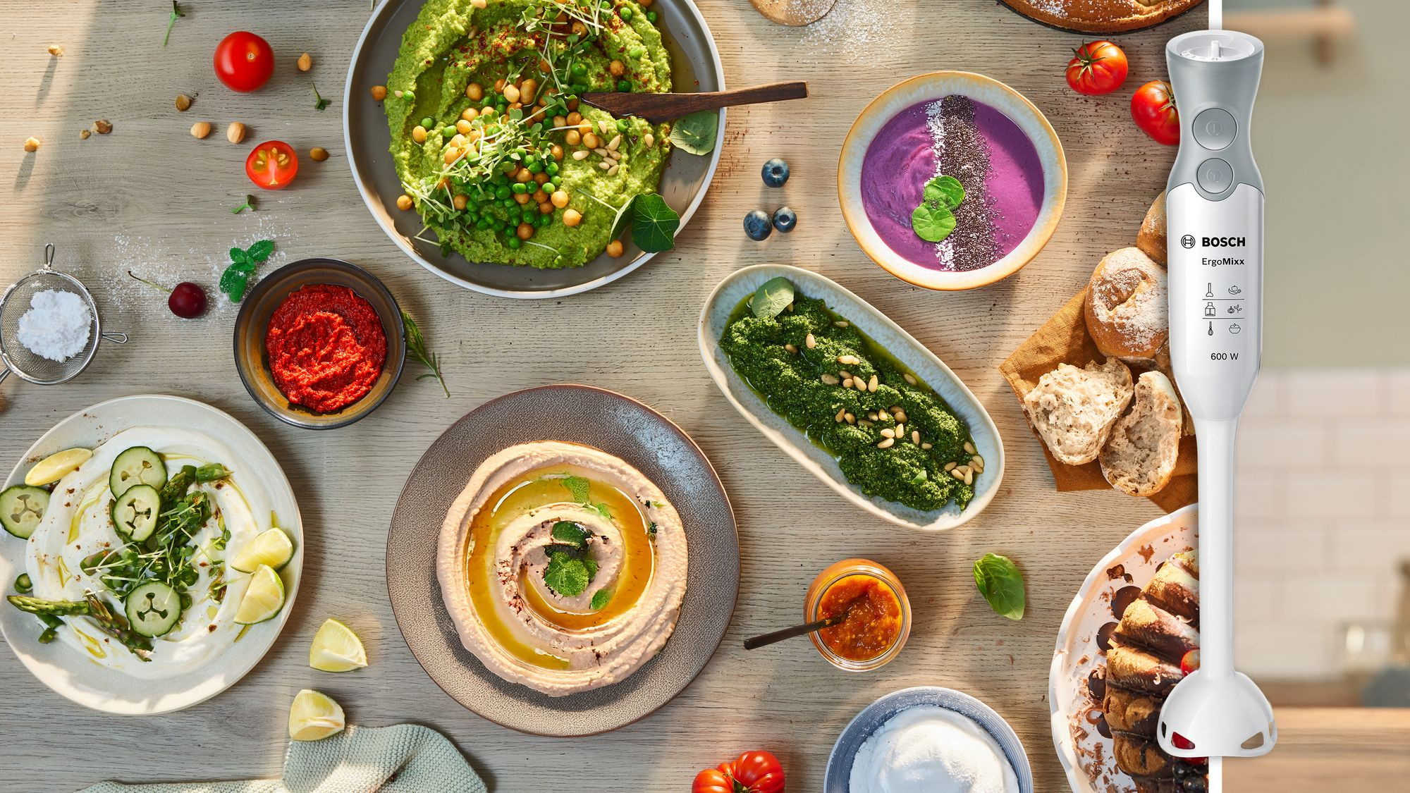 Ein Bosch-Stabmixer auf einem Holztisch, umgeben von Schüsseln mit Hummus, Pesto, Dips, Brot und frischen Zutaten.