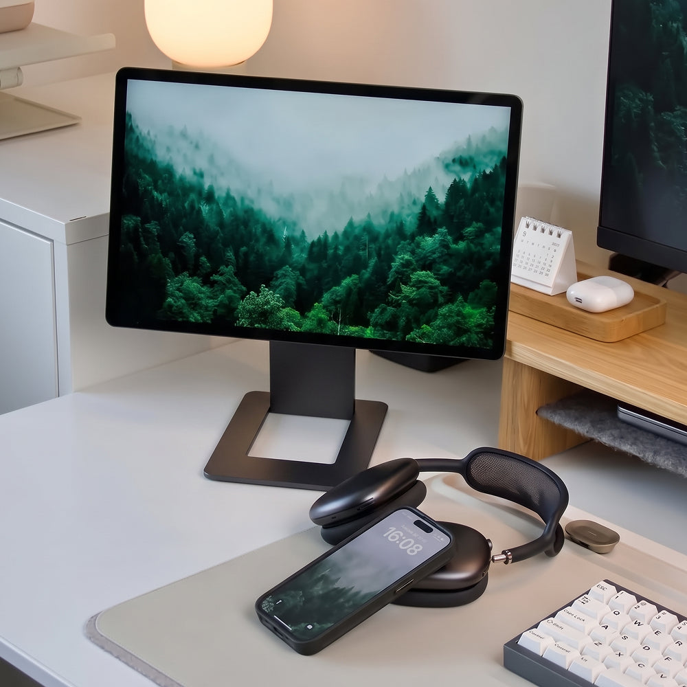 Un escritorio con un monitor que muestra un bosque, un teléfono, auriculares y un teclado blanco.