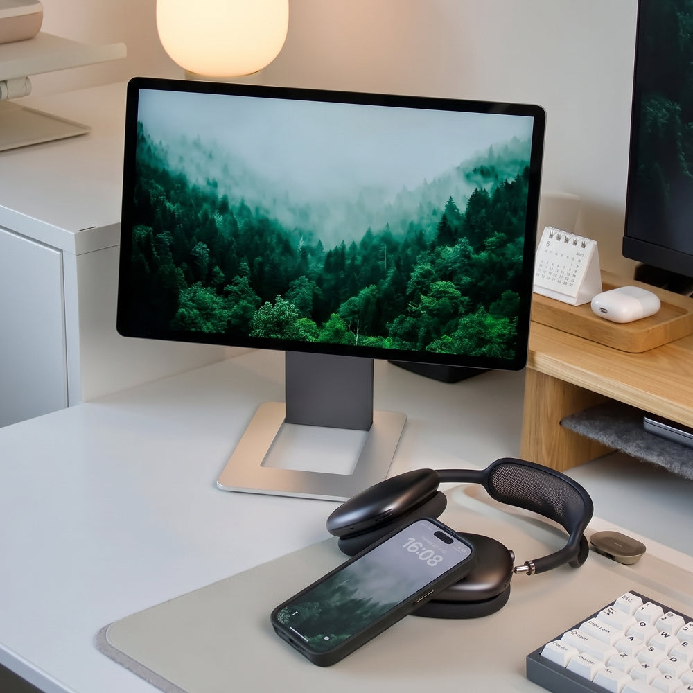 Un escritorio ordenado con un monitor con un bosque neblinoso, auriculares y un teléfono inteligente.