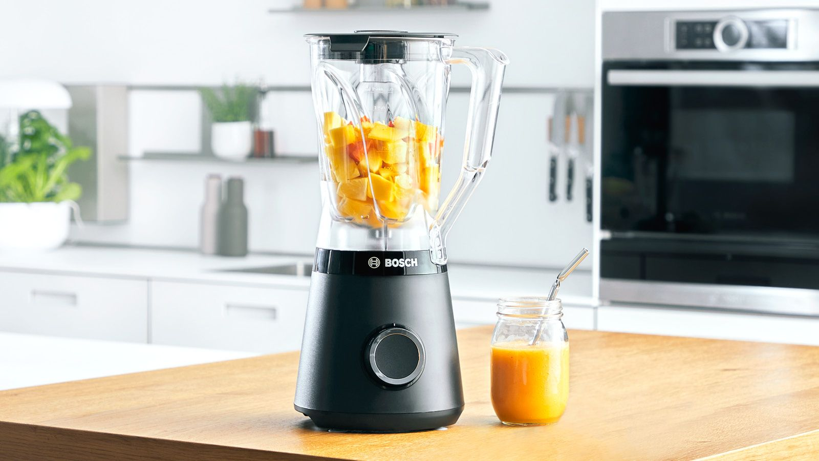 Un blender Bosch noir rempli de morceaux de fruits frais se trouve sur un comptoir en bois à côté d'un verre de smoothie.