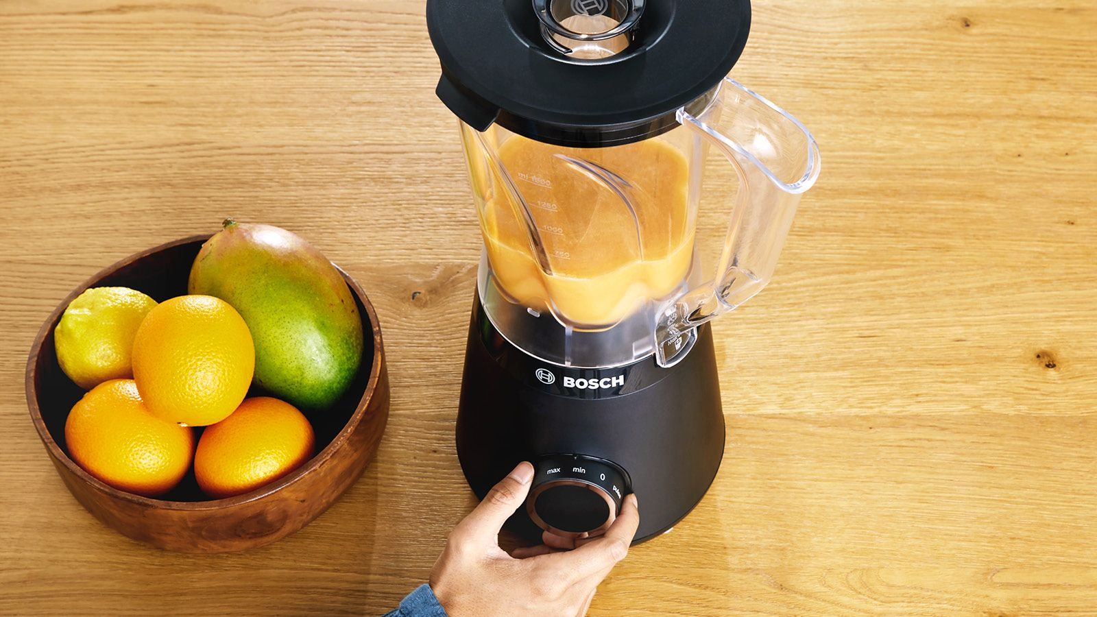 Une main actionne un blender Bosch noir avec un smoothie orange, à côté d'un bol de fruits frais.