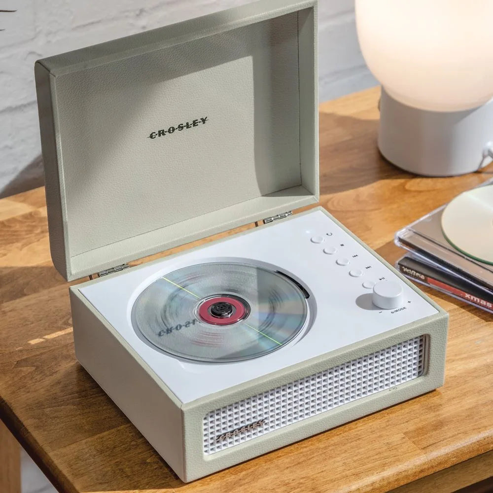 Ein cremefarbener Crosley-CD-Player mit offener Abdeckung steht auf einem Holztisch neben einer Lampe.