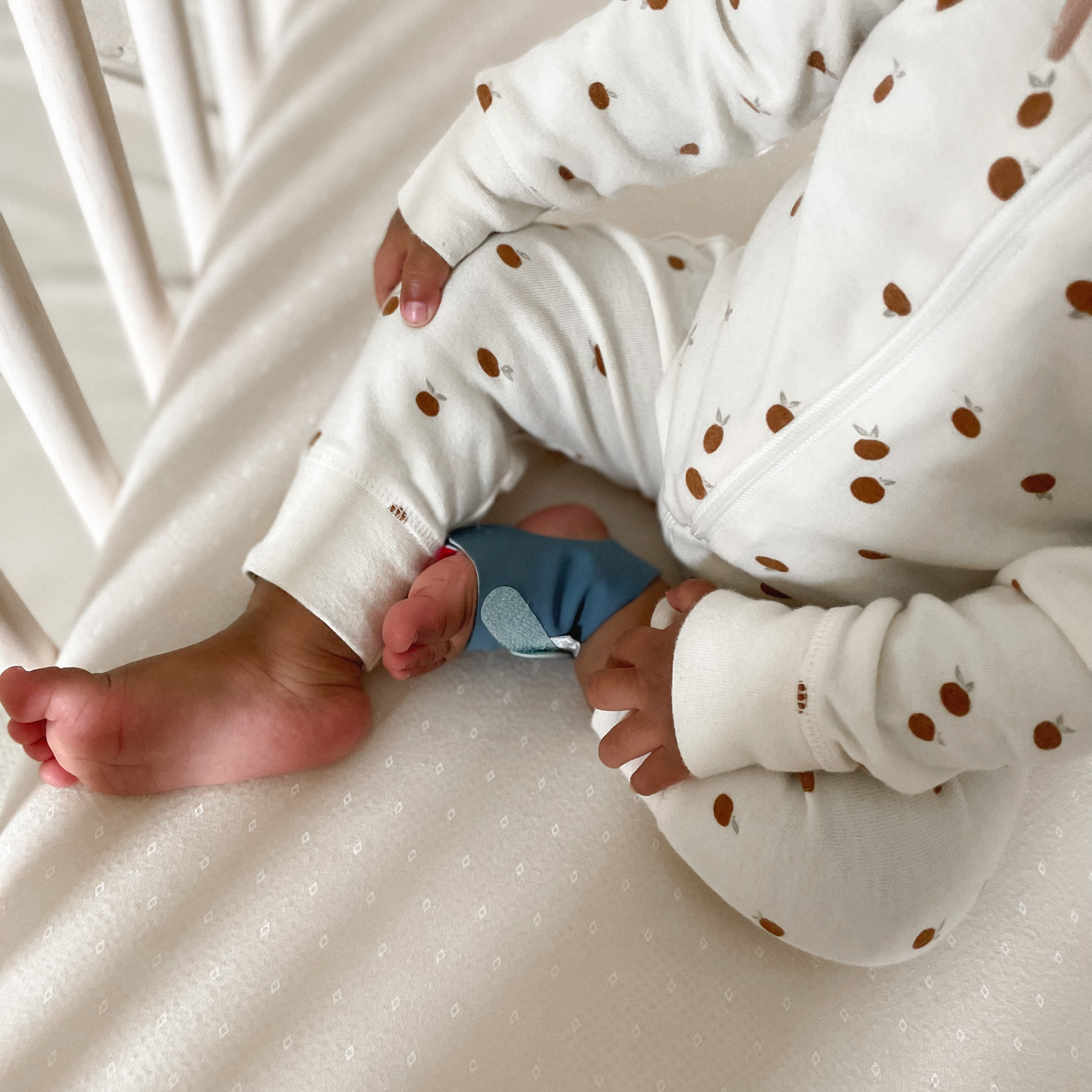 Baby in weißem Strampler mit Muster sitzt im Bett, ein Fuß trägt eine blaue Babymonitor-Socke.