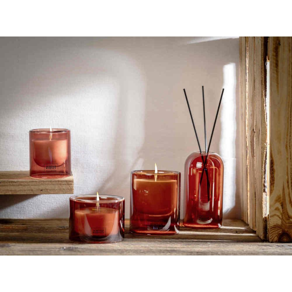 Tre candele profumate arancioni in barattoli di vetro e un diffusore con bastoncini su un ripiano.