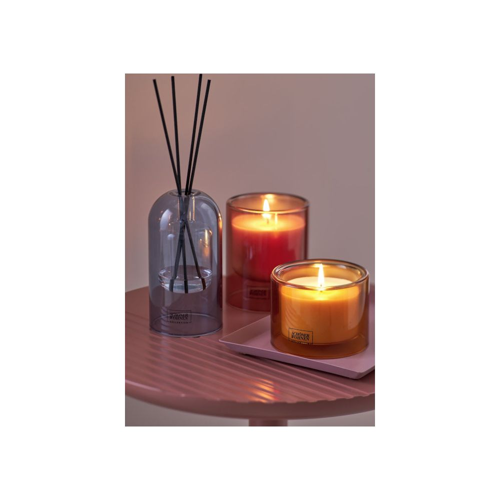 Due candele accese e un diffusore a bastoncini su un tavolo rotondo rosa contro una parete grigia.