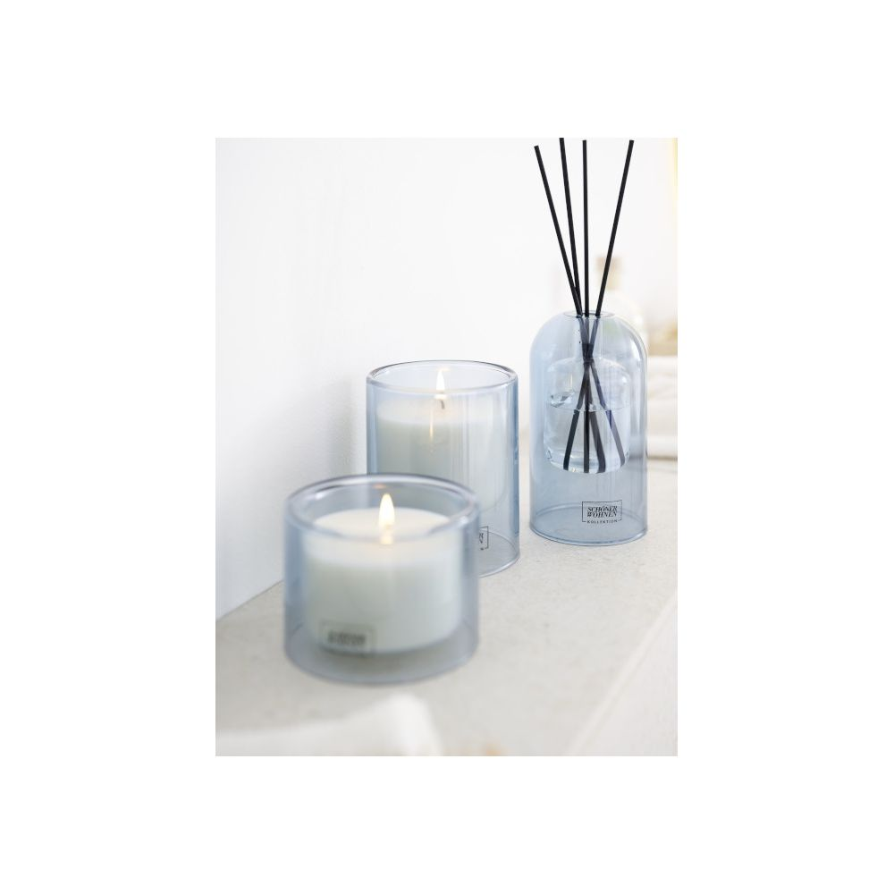 Due candele accese in bicchieri blu e un diffusore di fragranza poggiano su una superficie bianca.