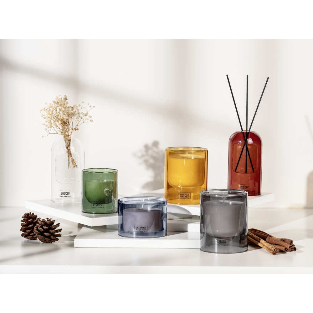 Diverse candele profumate e diffusori in barattoli di vetro colorato su supporti bianchi con pigne.