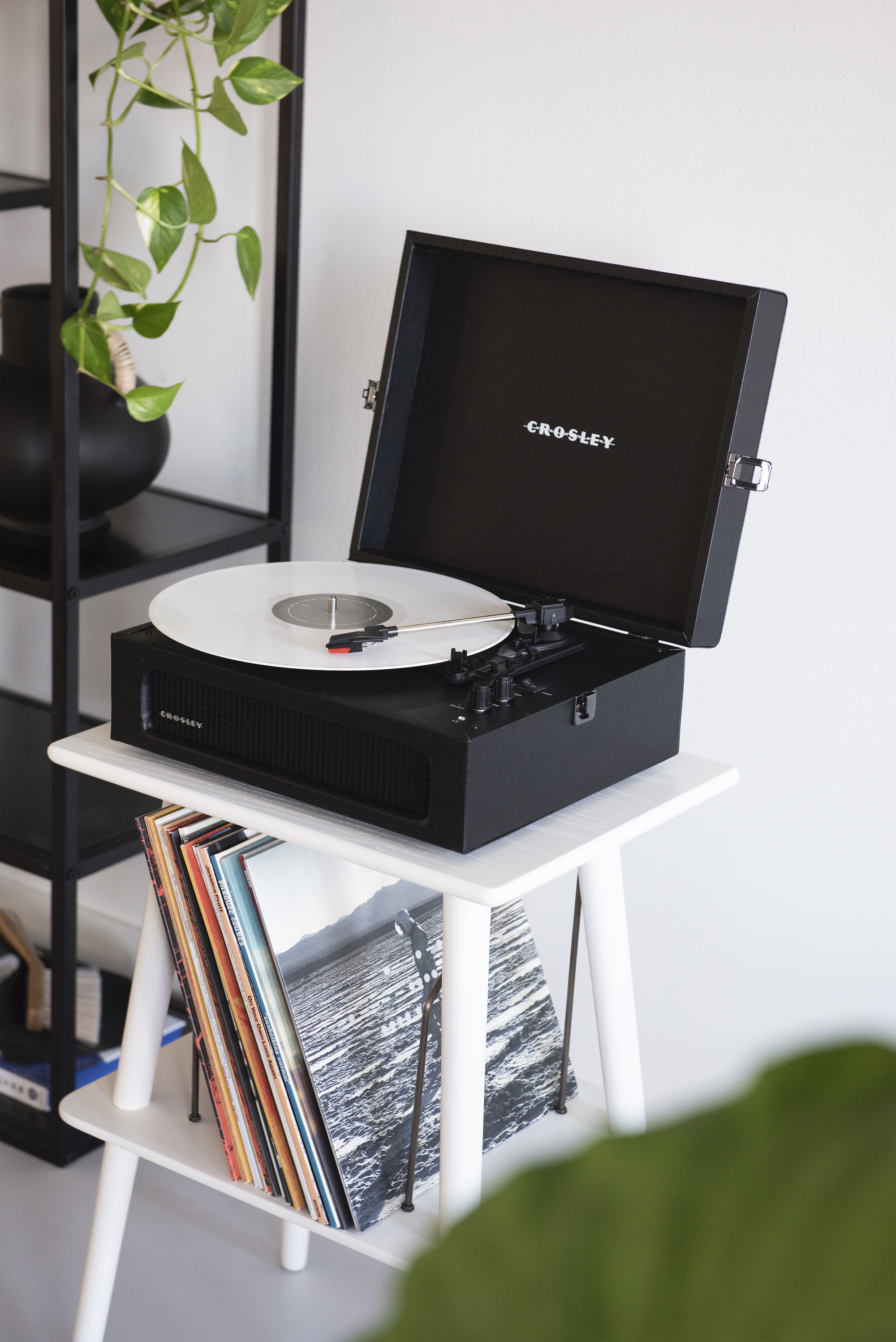 Ein schwarzer Crosley-Plattenspieler spielt eine weiße Platte auf einem weißen Tisch mit Schallplatten darunter.
