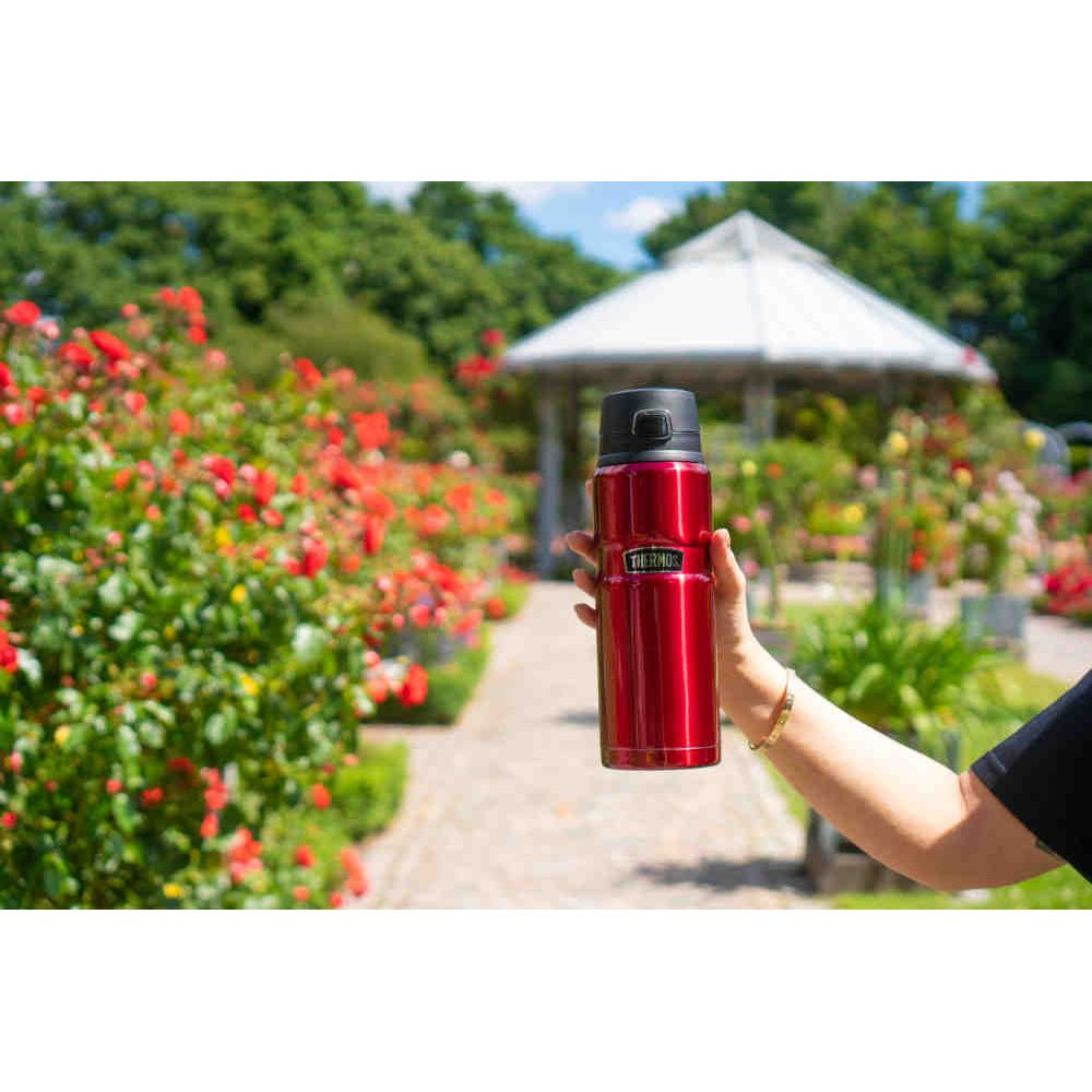 Eine Person hält eine glänzende rote Thermoskanne in einem Garten mit roten Blumen und einem Pavillon.