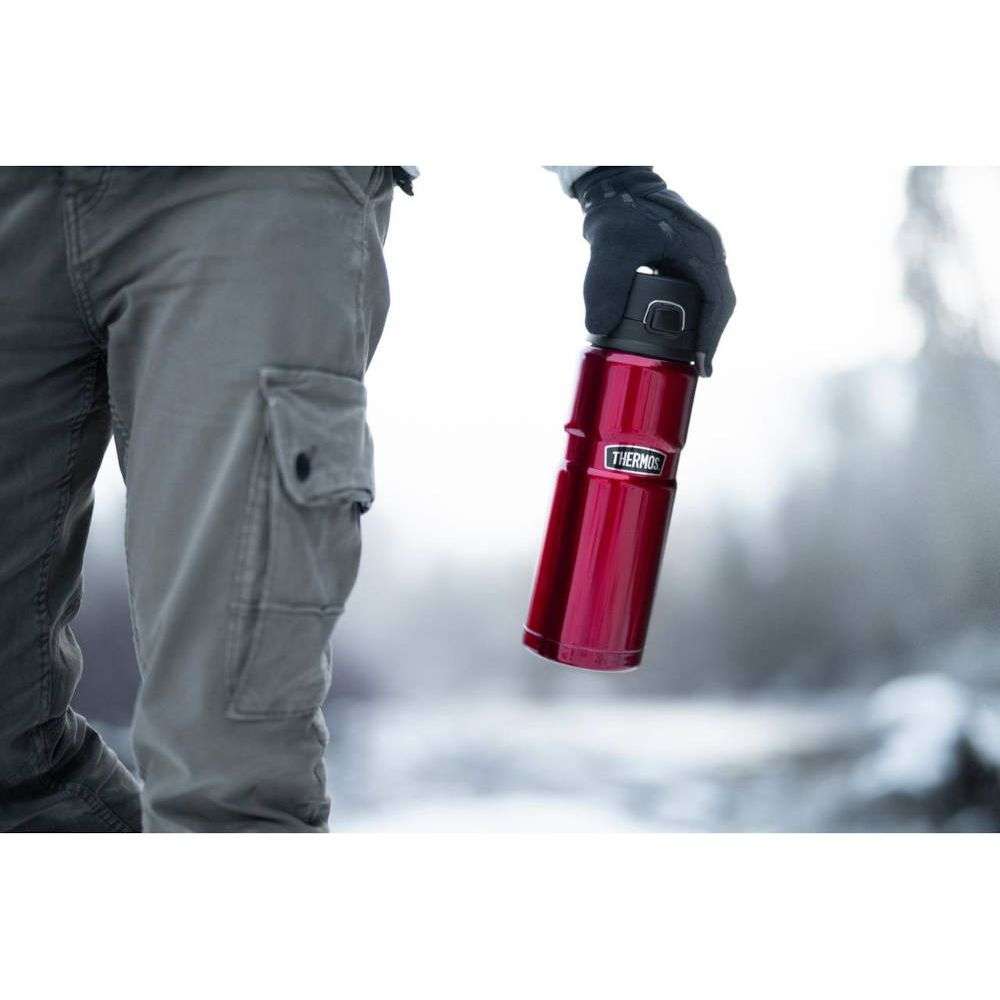 Eine Person im grauen Cargo-Hosen hält eine rote Thermosflasche in der verschneiten Natur.