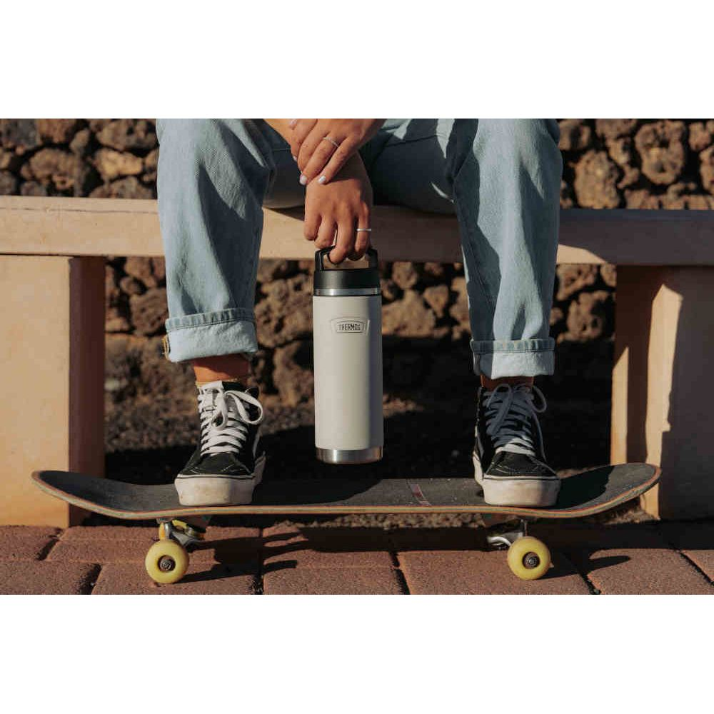 Person in Jeans und Sneakers hält eine Thermosflasche über ein Skateboard.