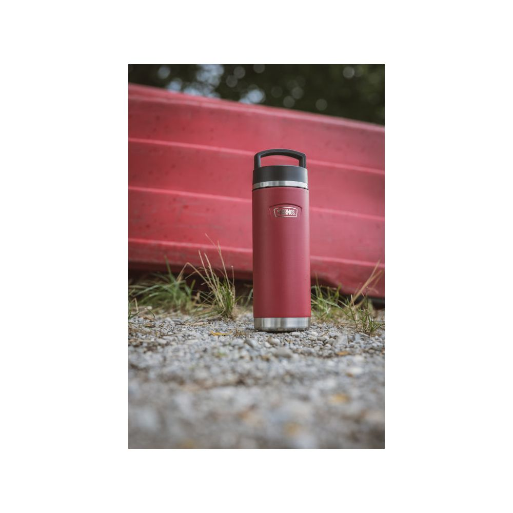 Eine rote Thermosflasche steht auf Schotter mit Gras, vor einem verschwommenen roten Boot und Bäumen.