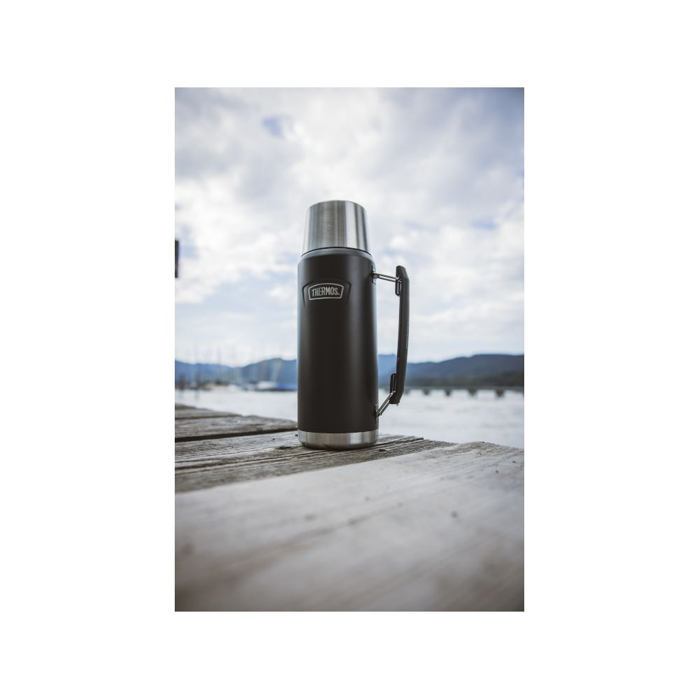 Eine schwarze Thermoskanne steht auf einem Holzsteg an einem Gewässer mit Bergen im Hintergrund.