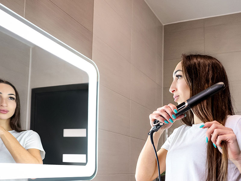 Una donna con lunghi capelli castani li liscia in un bagno piastrellato chiaro davanti a uno specchio illuminato.
