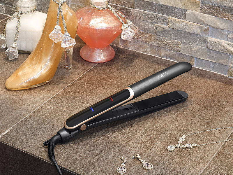 Una piastra per capelli nera e oro su una superficie di piastrelle marroni con flaconi di profumo e gioielli.
