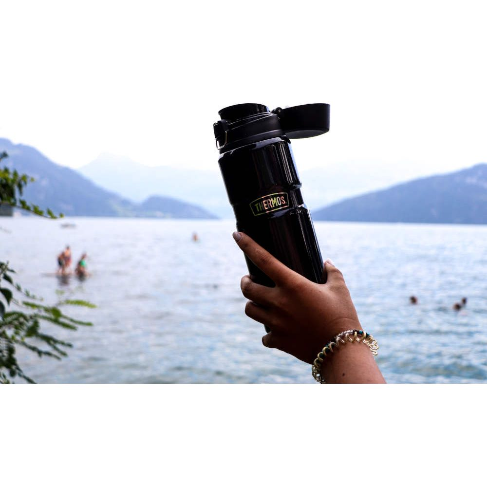 Eine Hand hält eine schwarze Thermoskanne vor einer See- und Bergkulisse.