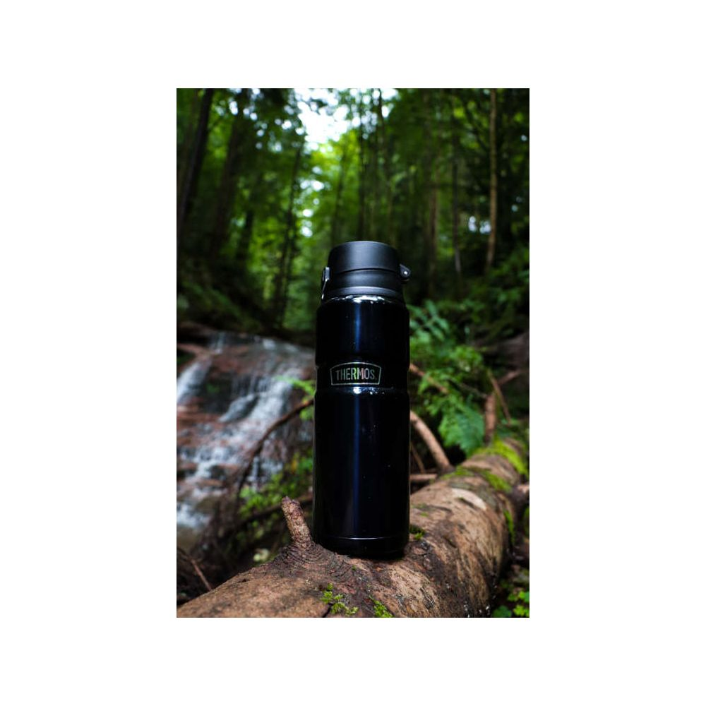 Eine schwarze Thermosflasche steht auf einem moosigen Baumstamm in einem grünen Wald mit verschwommenem Wasserfall.