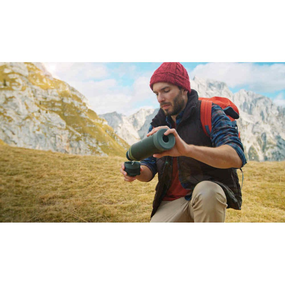 Ein Wanderer mit roter Mütze und Rucksack gießt Wasser aus einer Thermoskanne in eine Tasse vor verschneiten Bergen.
