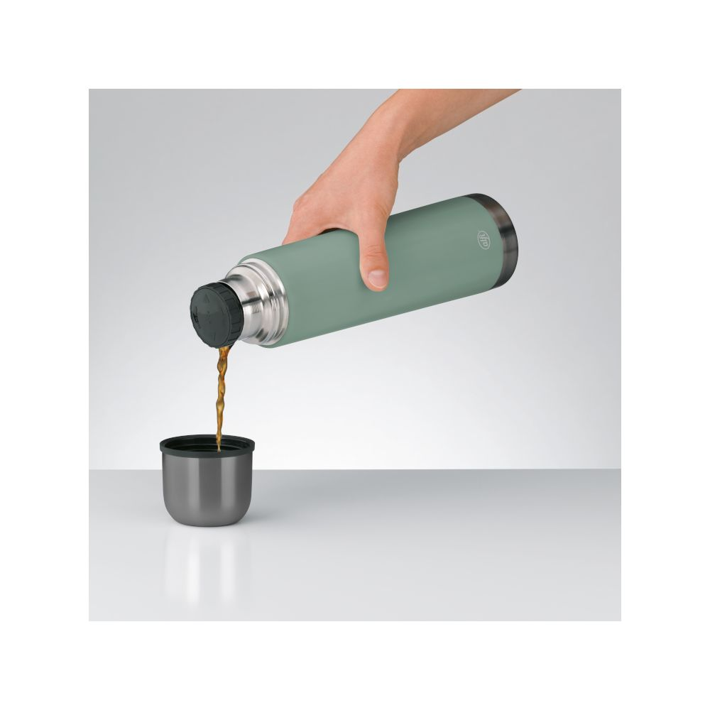 Eine Hand giesst Kaffee aus einer gruenen Thermoskanne in einen kleinen grau-schwarzen Becher.