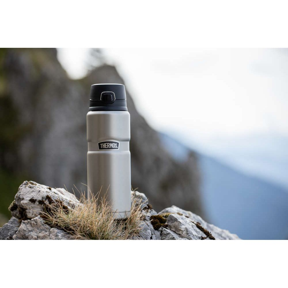 Eine silberne Thermoskanne steht auf einem felsigen Berggipfel mit Gras im Vordergrund.