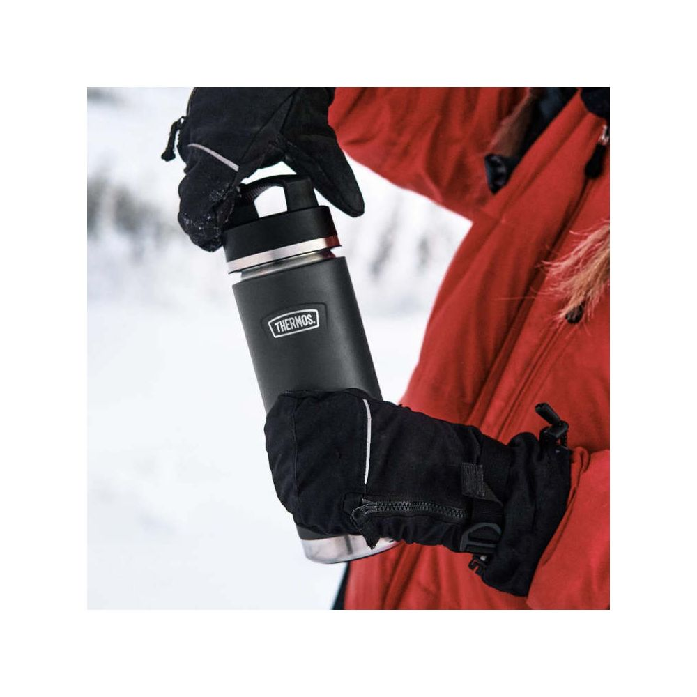 Eine Person im roten Mantel und mit schwarzen Handschuhen hält eine schwarze Thermoskanne im Schnee.