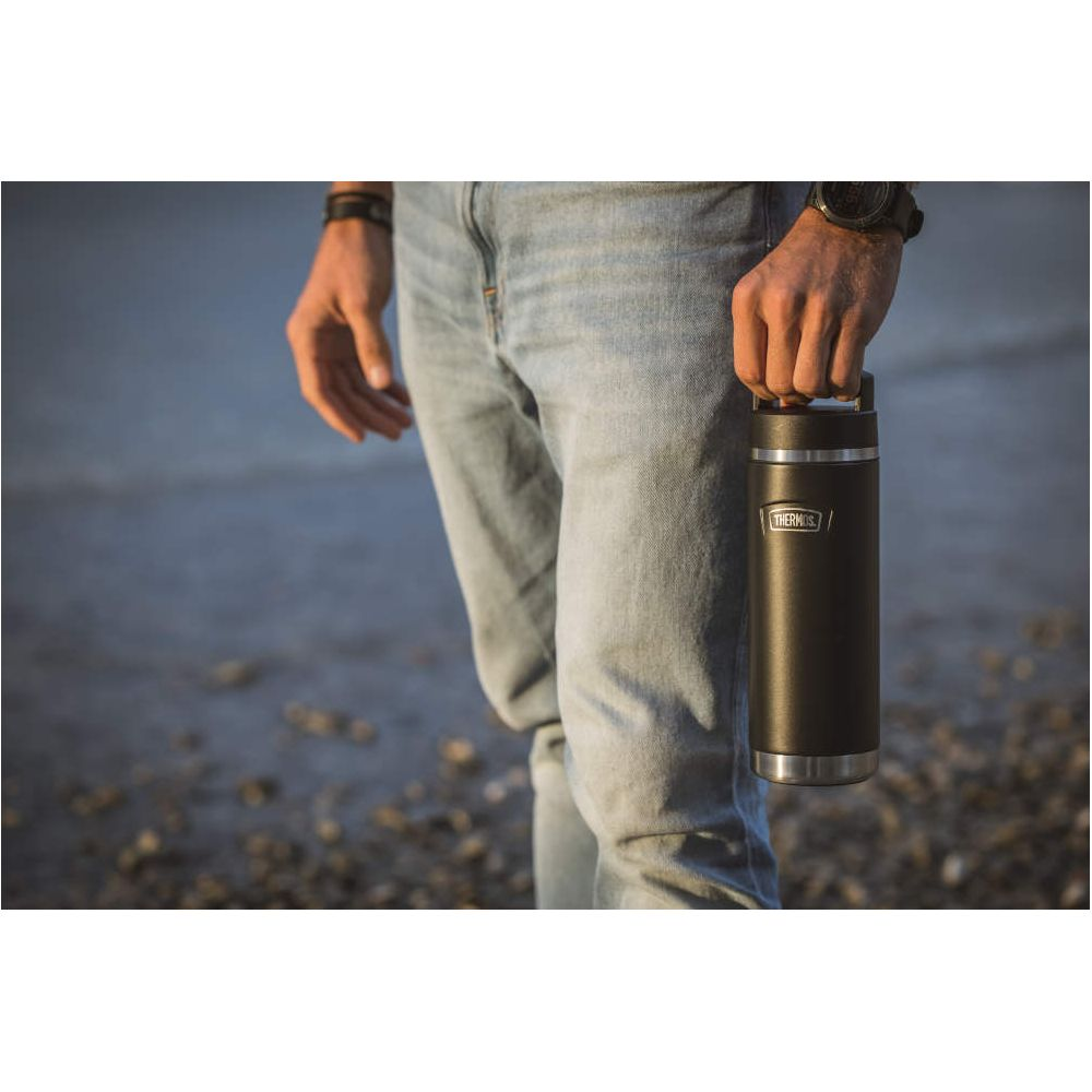 Eine Person in Jeans hält eine schwarze Thermoskanne an einem Strand.