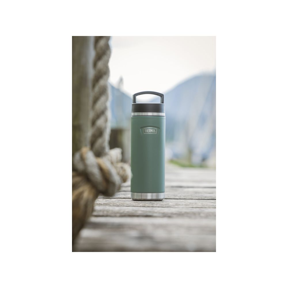 Eine grüne Thermosflasche steht auf einem Holzdeg mit Seil und verschwommenem Berg im Hintergrund.