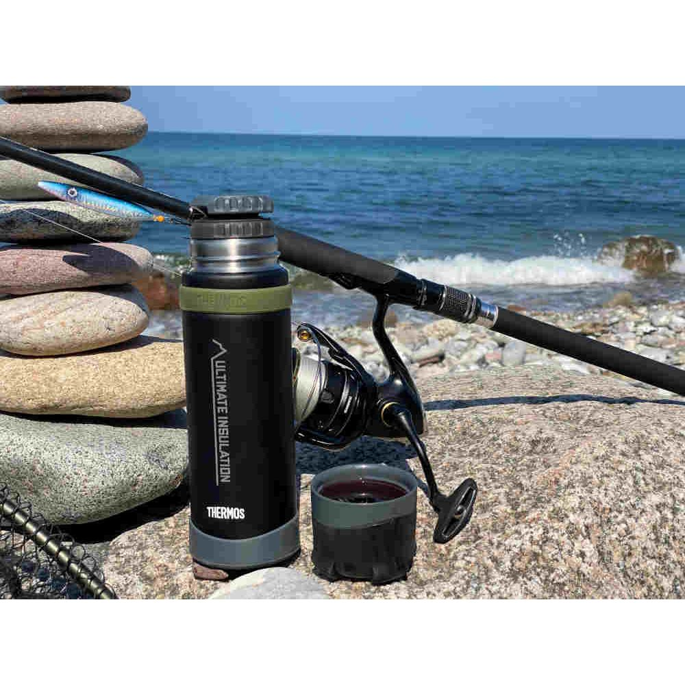 Eine schwarze Thermoskanne und eine Angelrute liegen auf felsigem Boden am Meer.